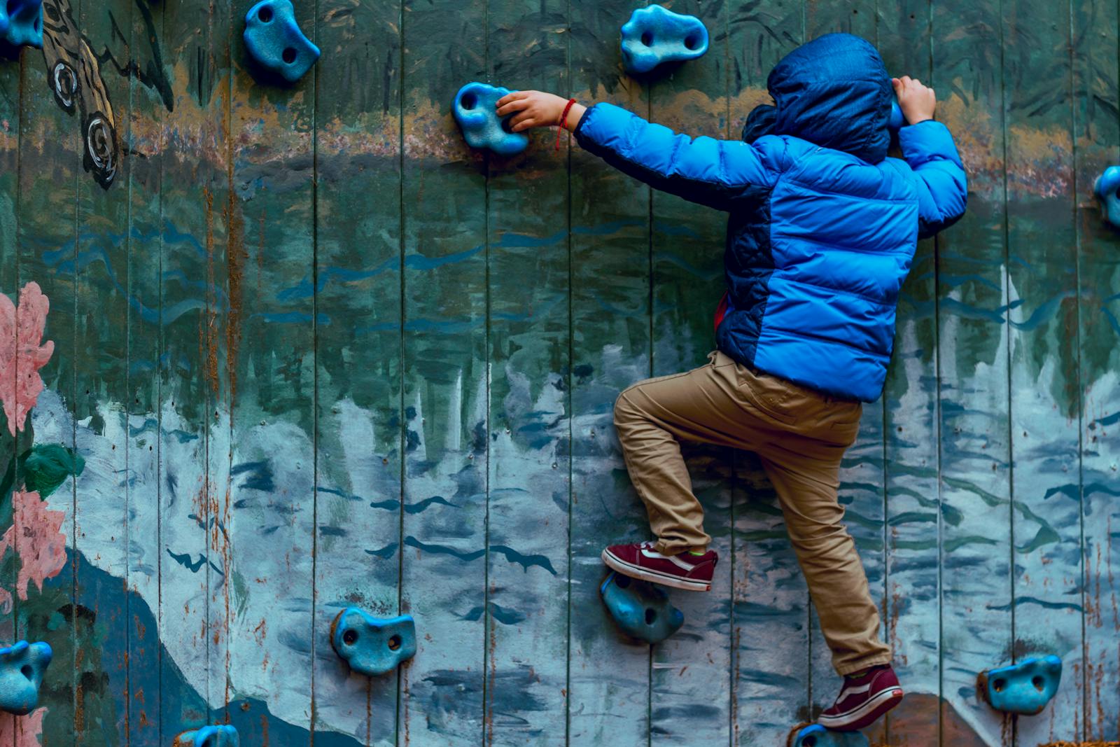 Young boy in blue jacket climbing a painted rock wall outdoors, showcasing adventure and play.